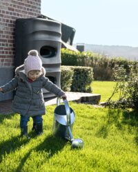 klein kind met gieter die gras water geeft met water uit de regenton op de achtergrond tegen het huis