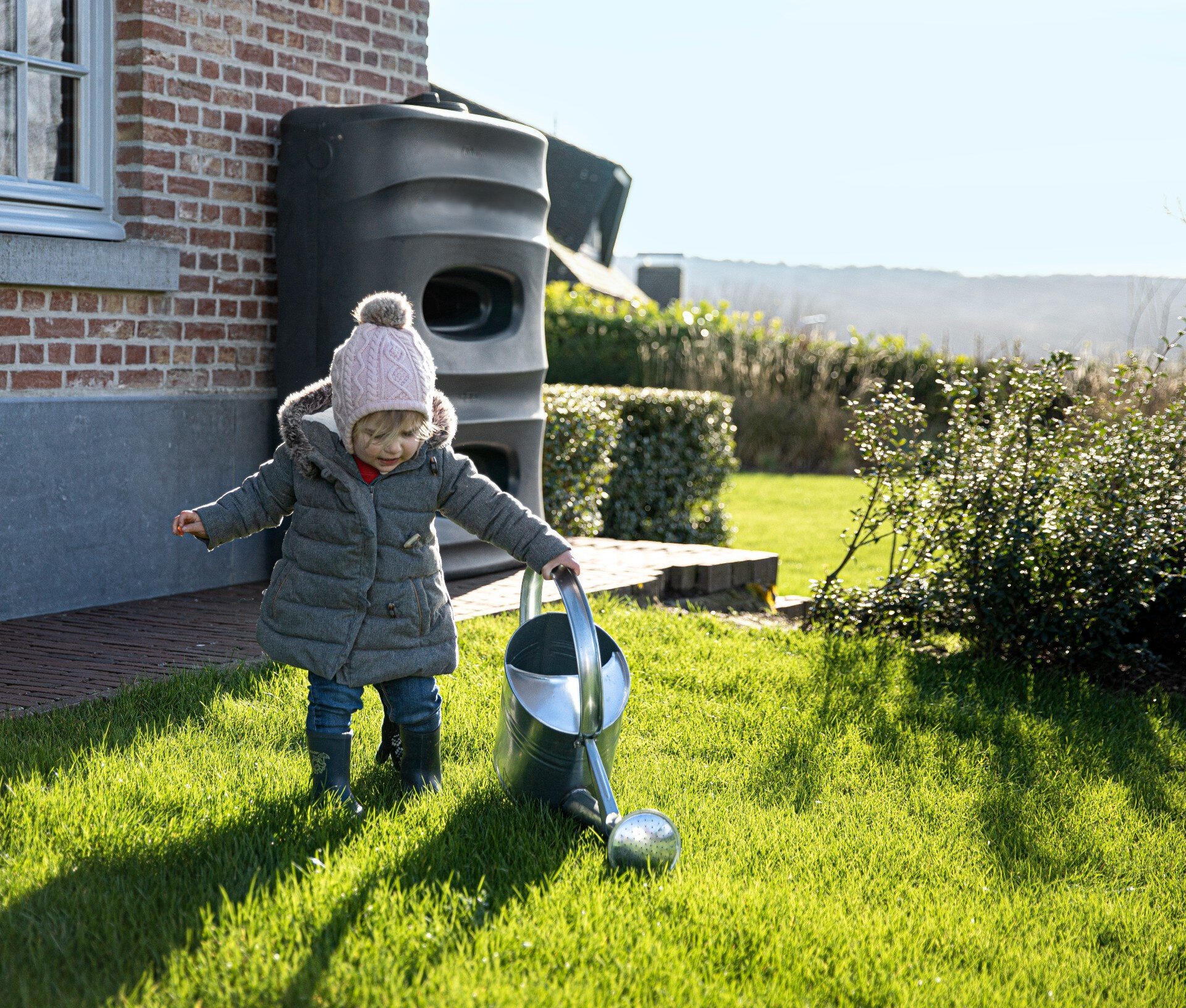 klein kind met gieter die gras water geeft met water uit de regenton op de achtergrond tegen het huis